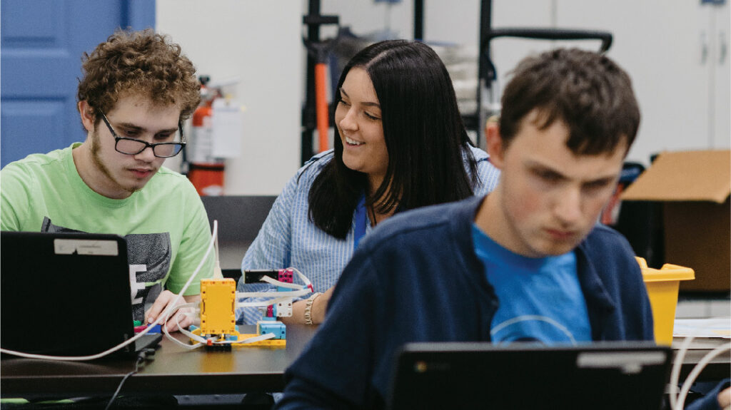 a woman smiling at a young man as they work on a building a small robot at a table