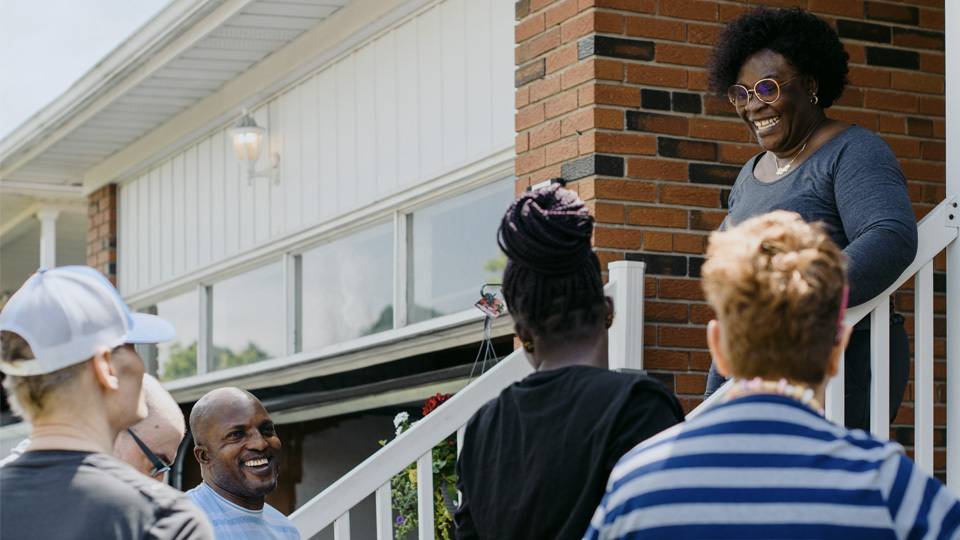 A group of people standing in front of a house. One woman stands on the porch looking down at them and smiling.